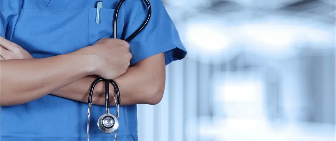 A nurse in scrubs with arms crossed, holding a stethoscope in a hospital corridor