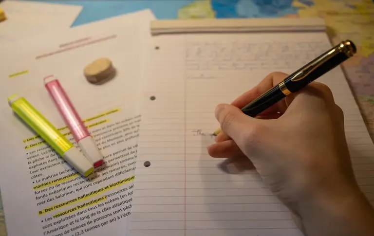 A hand writing notes beside highlighters and a printed article